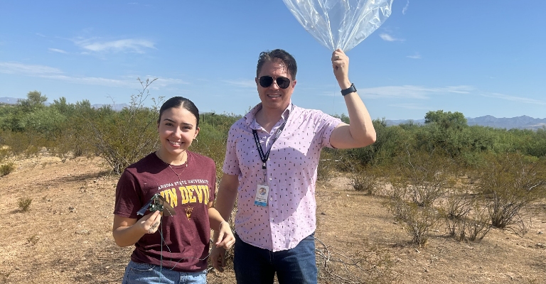 A woman holds an electronic device next to a man holding a weather balloon in the desert