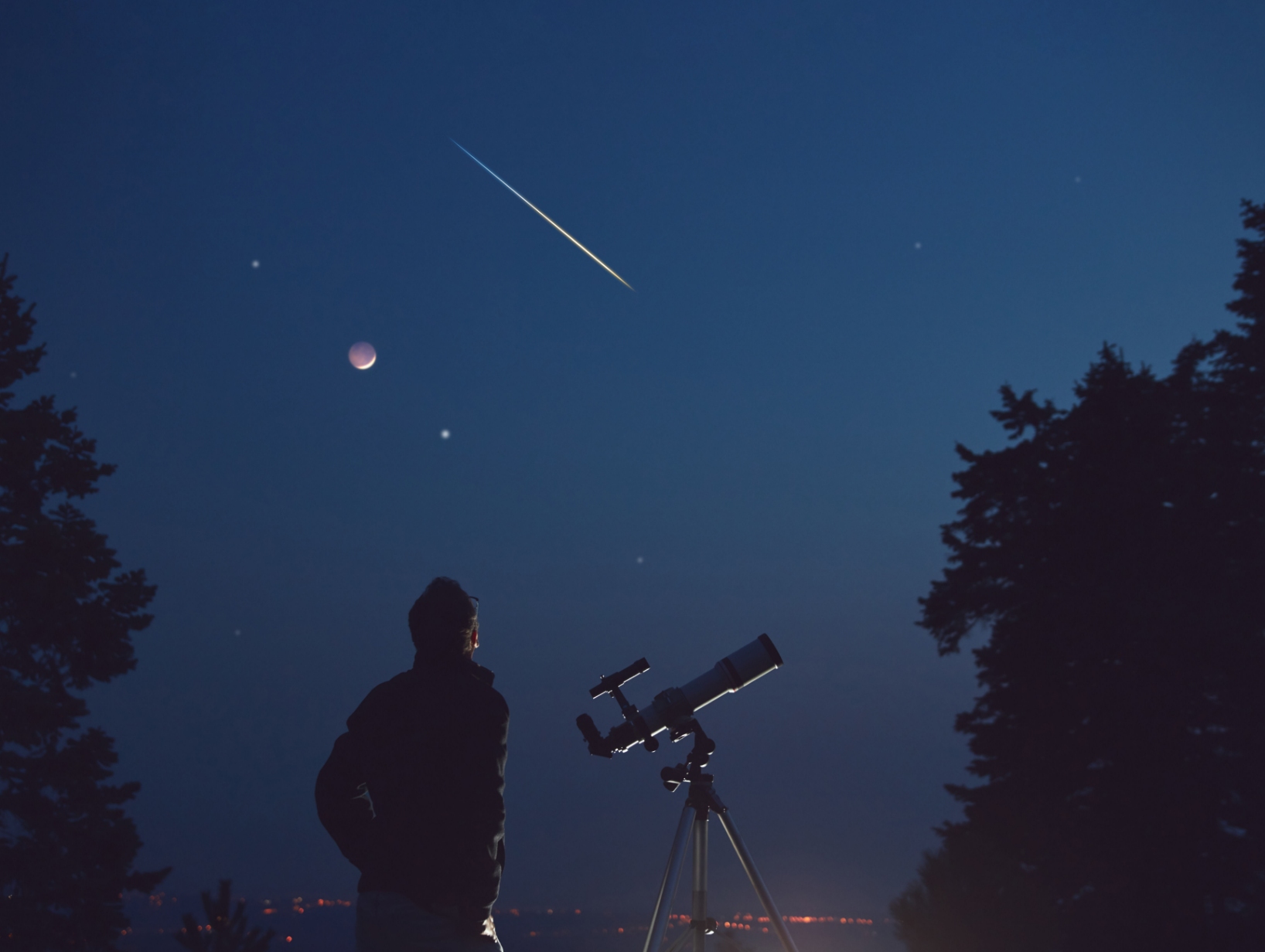 A man stands next to a telescope, looking up at planets and a meteor in the night sky